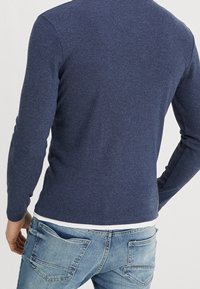 Homme portant une chemise à manches longues bleu marine par-dessus un t-shirt blanc et un jean en denim bleu, vu de dos sur un fond uni.
