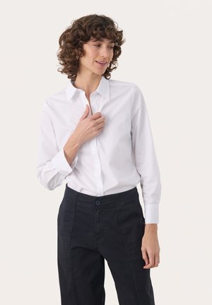 Femme aux cheveux bouclés portant une chemise blanche boutonnée et un pantalon noir, ajustant le col de sa chemise, regardant légèrement vers la gauche.