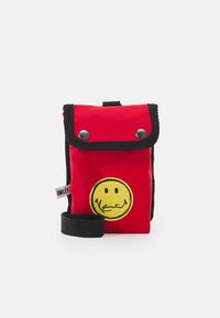 Röd tygväska med svarta accenter, tryckknappsstängning och design av en gul smiley. Har en axelrem och en tagg på sidan.