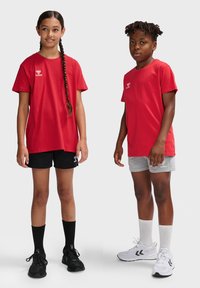 Røde bomulds T-shirts med logo, parret med sorte og grå shorts. Begge har sportssko og sokker på og står mod en neutral baggrund.