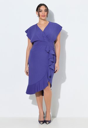 Kvinde iført en lilla slå-om-kjole med flæsedetaljer på nederdelens forside og kapærmer, kombineret med sorte åbne hæle.