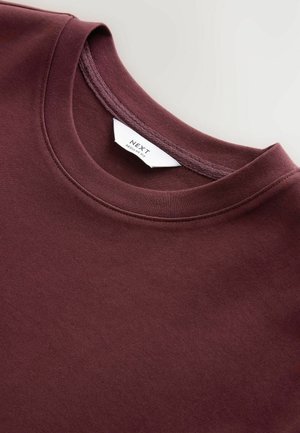 Sweatshirt bordeaux à col rond fabriqué en mélange de coton doux, doté d'une texture lisse, d'une coupe régulière et d'une étiquette de marque visible sur le col.