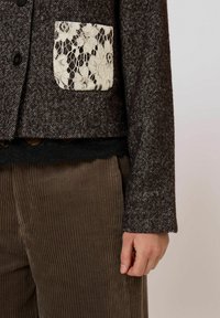 Hnědý kabát s vzorem psí zub, s bílou krajkovou kapsou a černým ozdobným lemováním, doplněný texturovanými hnědými manšestrovými kalhotami.