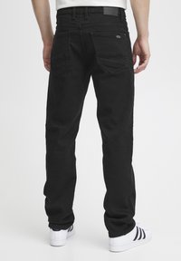 Black denim jeans with a straight cut, visible stitching, two back pockets, and a label at the waistband. Worn with white sneakers.