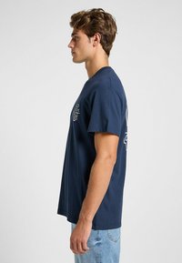 Camiseta de algodón azul marino de manga corta, con cuello redondo y diseño gráfico en la parte frontal, que presenta un logo y un año. Jeans de color azul claro.