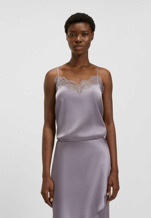 Donna di carnagione scura che indossa un top in raso lilla con pizzo e una gonna coordinata, in piedi davanti a uno sfondo chiaro e neutro.