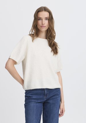 Femme aux longs cheveux ondulés portant un pull blanc à manches courtes et un jean bleu, debout, une main sur la hanche, devant un fond blanc.