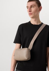 Beige schoudertas van soepel leer, met een ritsluiting en een brede stoffen schouderband, gedragen door een model in een zwart shirt.