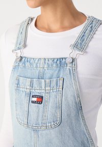 Šviesiai mėlynos džinsinės kombinezonas su sidabriniais priedais, su reguliuojamomis petnešomis, priekinėmis kišenėmis ir "TOMMY JEANS" žyme. Minkšta tekstūra.