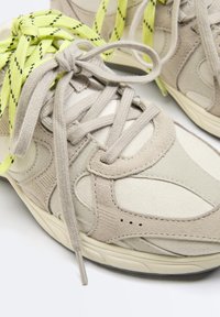 Baskets en daim beige et tissu avec des lacets épais beiges et des lacets contrastants jaunes et noirs en arrière-plan.