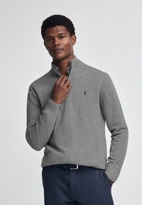 Pullover grigio a maglia con colletto zip, polsini a coste e un piccolo logo ricamato sul petto. Indossato sopra una camicia a righe.