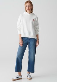 Felpa bianca in cotone con tasca a marsupio frontale, vestibilità casual, abbinata a jeans blu chiaro a gamba dritta e sneakers bianche con strisce blu navy.