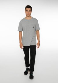 Grijze korte mouwen t-shirt met een borstzak en zwart logo, gecombineerd met zwarte broek en zwarte sneakers met gekleurde accenten.