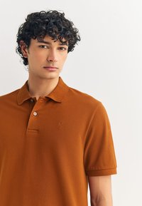 Ruda textura audinio polo marškinėliai su klasikiniu apykakliu, dviem mygtukais ir mažu siuvinėtu logotipu ant krūtinės.