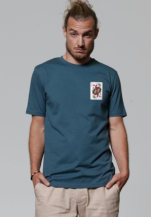 Man met gebonden haar die een teal t-shirt draagt met een afbeelding van de Koningin van Harten speelkaart op de borst, staand met handen in de zakken.