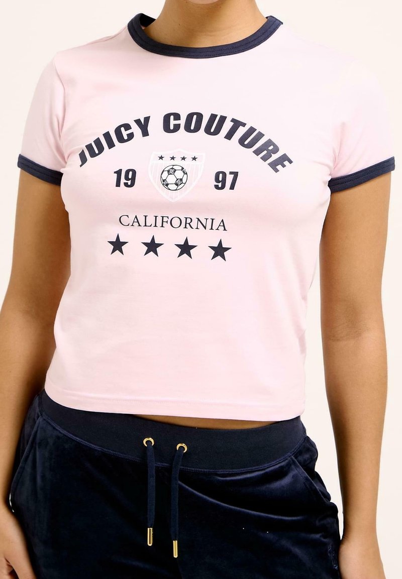 Světle růžové zkrácené tričko s námořnickým modrým límečkem a rukávy, na němž se nachází nápis "Juicy Couture," rok "1997" a fotbalová grafika v námořnické.