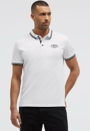 Man wearing a white polo shirt with black trim on collar and sleeves, and black text logo on chest, standing against plain background.