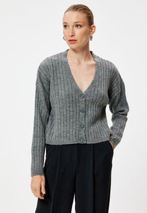 Grijze geribbelde cardigan met een diepe V-hals, voorzien van vijf knoopsluitingen en lange mouwen, gecombineerd met donkere, op maat gemaakte broeken.