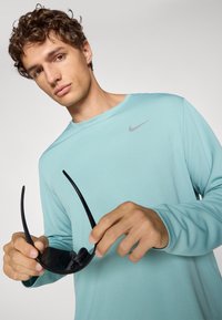 Lyseblå langærmet skjorte med et gråt Nike-logo, holdt af en person, der griber fat i sorte solbriller, med en glat stofstruktur.
