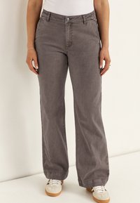 Person iført løstsiddende, falmede grå bukser med frontknapper, lommer og hvide sneakers, stående mod en ensfarvet baggrund.