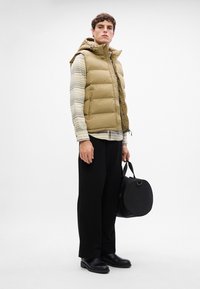 Gilet imbottito beige con cappuccio, tasche con zip, indossato sopra una camicia a righe. Pantaloni neri a gamba larga e stivali neri lucidi. Borsa rotonda nera.