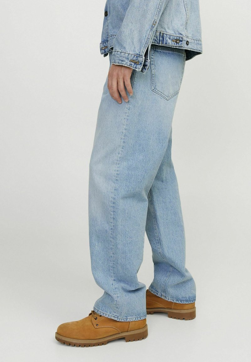 Vaqueros de mezclilla de color azul claro con un corte relajado, costuras visibles y bolsillos laterales, combinados con botas de trabajo de color marrón claro. La mezclilla presenta una textura ligeramente desgastada.