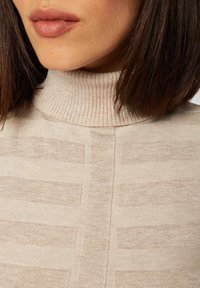 Beżowy sweter z golfem ma prążkowaną strukturę i poziome paski. Materiał jest miękki z delikatnym połyskiem, podkreślający elegancki fason.