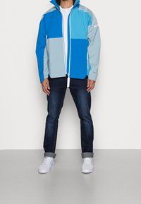 Muž oblečený v modro-modrošedém barevném zipovém kabátu, bílém tričku, tmavých vykasaných džínách a bílých teniskách, stojící proti bílé zdi.