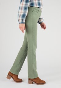 Pantaloni di cotone verdi con una vestibilità slim, caratterizzati da una texture liscia e cuciture sottili, abbinati a stivaletti con tacco in suede marrone.