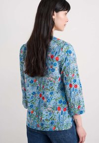 Blouse bleue à motif floral avec des manches trois-quarts. Fabriquée en tissu léger, elle présente des motifs botaniques rouges, bleus et verts.