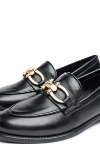 Black leather loafers featuring a gold circular buckle detail on the front and stitched accents around the edge. Classic low-profile design.