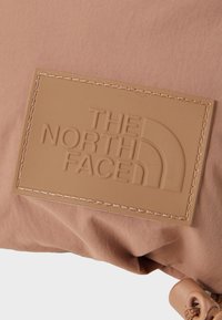 Pink fabric bag featuring a tan rubber logo patch with embossed text. Includes stitching details and a cord lock closure.