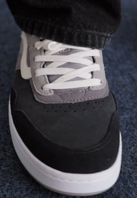 Gray and black sneaker with a suede and fabric upper, white laces, and a white rubber sole. Features perforations on the toe.