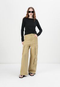 Zwarte langemouwen top gecombineerd met beige cargobroek met klepzakken, een relaxed fit en platte sandalen, tegen een effen witte achtergrond.