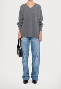 Pull en maille grise avec col en V, coupe ample et texture côtelée ; jean bleu avec un design graphique subtil ; tenant un sac à main noir.