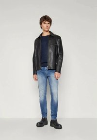 Fekete bőr dzseki cipzárral, alatta kék pulóver, viseltes világoskék farmer, és fekete bokacsizma. Semleges háttér előtt áll.