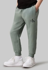 Pantaloni jogger verde menta con polsini a costine e tasche laterali, con un logo nero sulla coscia. Indossati con sneakers bianche.