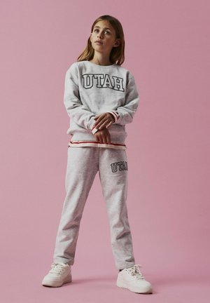 Szara bluza i dresy z napisem "UTAH" w ciemnych literach. Czerwono-białe paski na mankietach. Białe sneakersy. Jednolity różowy tło.