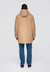 Beige huvad kappa med enkel design, med långa ärmar och rak skärning, kombinerad med blåa denimjeans och mörka skor.