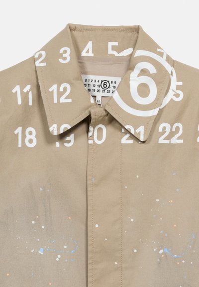 Μπεζ πουκάμισο με γιακά, με λευκούς αριθμούς και πιτσιλιές από μπογιά τυπωμένες, που διαθέτει ετικέτα με έναν κυκλωμένο αριθμό έξι μέσα στον γιακά.