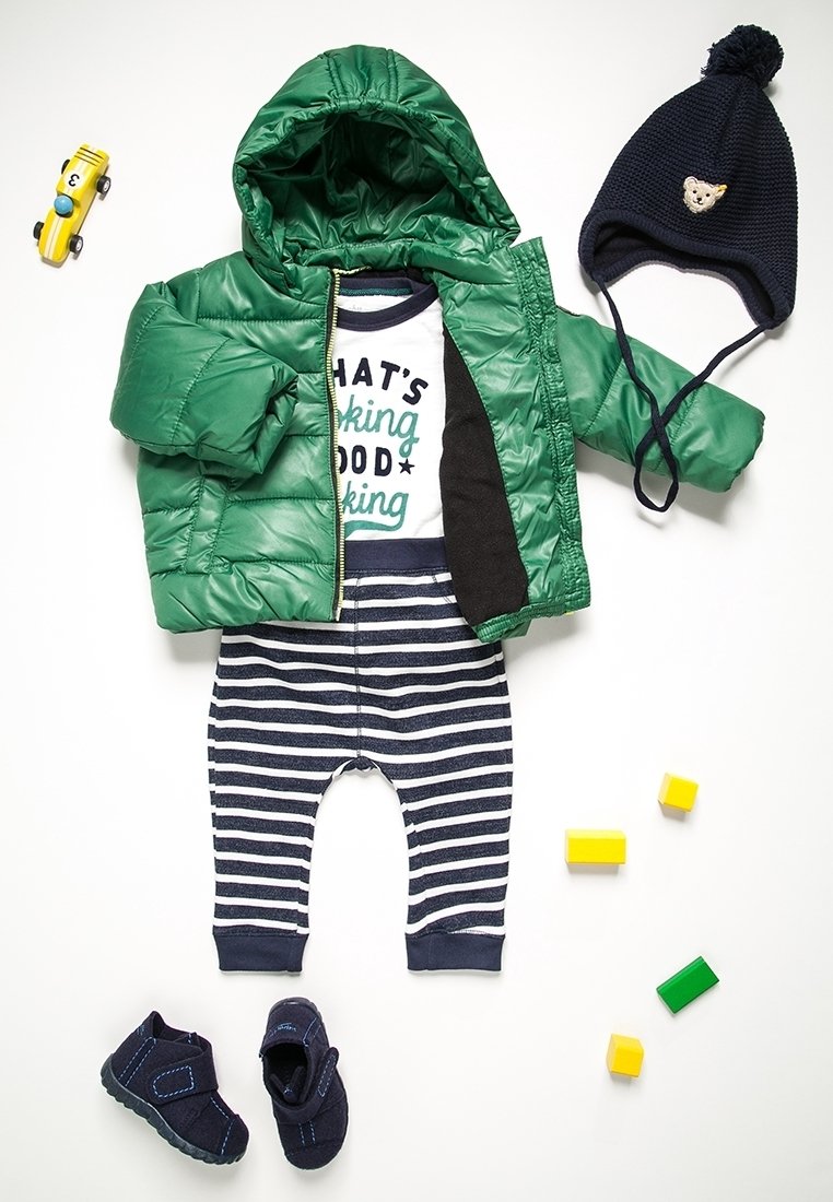 Zelená nadýchaná bunda, námořnický pruhovaný overálek s nápisem, námořnické botičky, modrá pletená čepice s medvídkem, žluté a zelené hračky v podobě kostek poházené kolem.