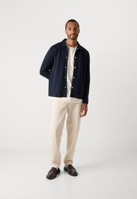 Man standing wearing a navy buttoned jacket over a white shirt, cream pants, and dark brown loafers on a plain light background.