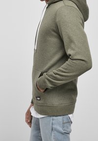 Ljus olivgrön hoodie med dragsko i huvan, framtill ficka och ribbade ärmslut och fåll. Bärs över en vit skjorta med en liten logotyp patch.