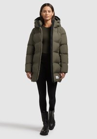 Olivově zelená péřová bunda s kapucí, předním zipem a bočními kapsami. Černé akcenty podél zipu. Nošena přes tmavý top a černé legíny.
