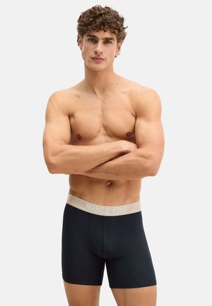 Muskulöser Mann mit lockigem Haar, der schwarze Marc O'Polo Boxershorts trägt und mit verschränkten Armen vor einem einfarbigen Hintergrund steht.