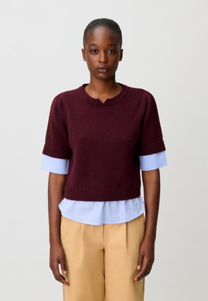 Giovane donna con i capelli corti che indossa un maglione bordeaux a maniche corte sopra una camicia a righe blu e pantaloni beige, in piedi davanti a uno sfondo uniforme.