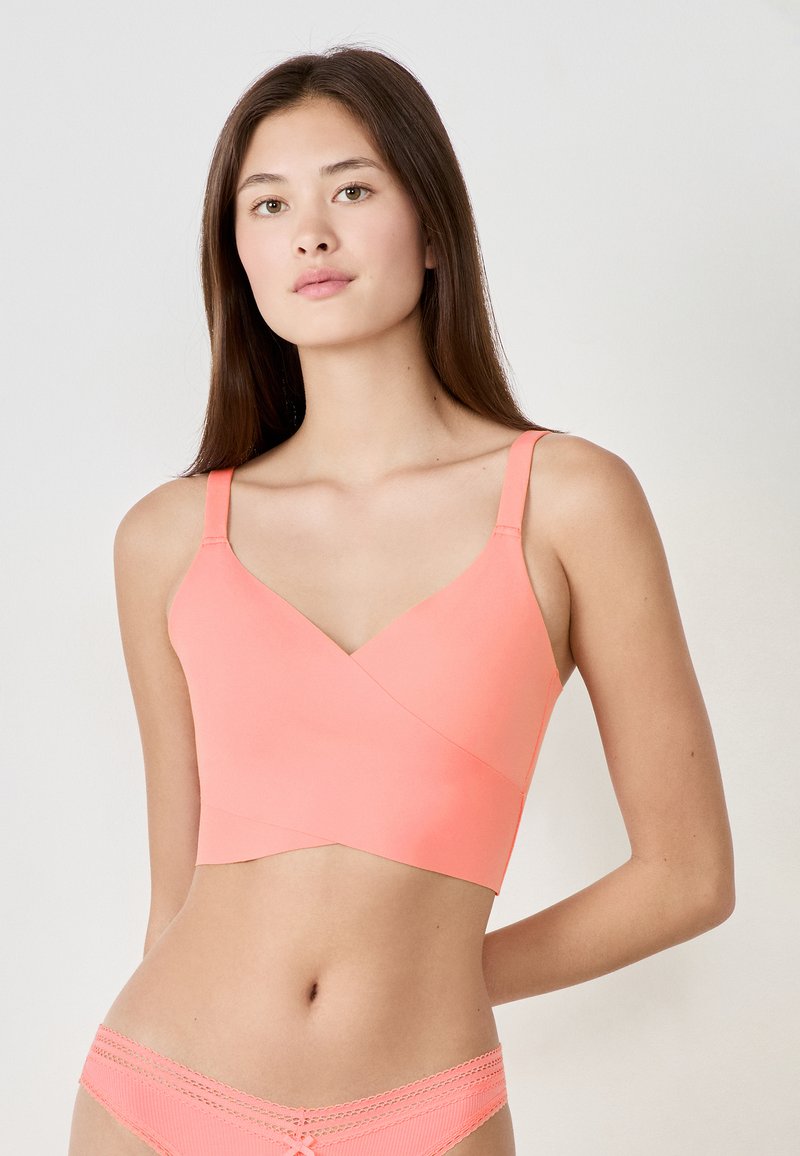 Koronkowy biustonosz w kolorze koralowym z krzyżowym fasonem, wykonany z gładkiego materiału. Cienkie ramiączka i dopasowany kształt, w zestawieniu z pasującym wykończeniem bielizny.