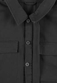 Czarna koszula zapinana na guziki z klasycznym kołnierzykiem, dwiema kieszeniami na piersi oraz trwałym materiałem o teksturowanym wykończeniu. Guziki są czarne i okrągłe.