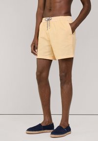 Shorts de bain jaunes à rayures avec une taille élastique et un cordon de serrage, dotés de poches latérales. Portés avec des espadrilles marines.