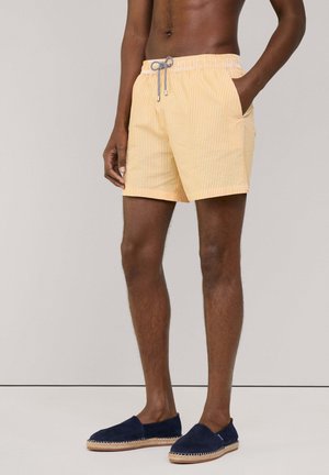 Gelbe Streifen-Badeshorts mit einem elastischen Bund und Kordelzug, ausgestattet mit seitlichen Taschen. Getragen mit marineblauen Espadrilles.
